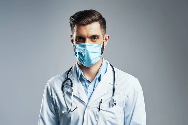 Confident young man in white lab coat and protective mask looking at camera while standing against grey background — Stock Photo, Image