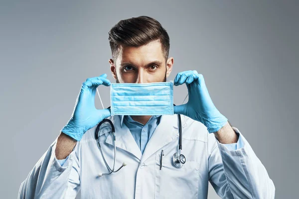 Hombre joven y seguro en bata de laboratorio blanca ajustando la máscara protectora mientras está de pie sobre fondo gris — Foto de Stock