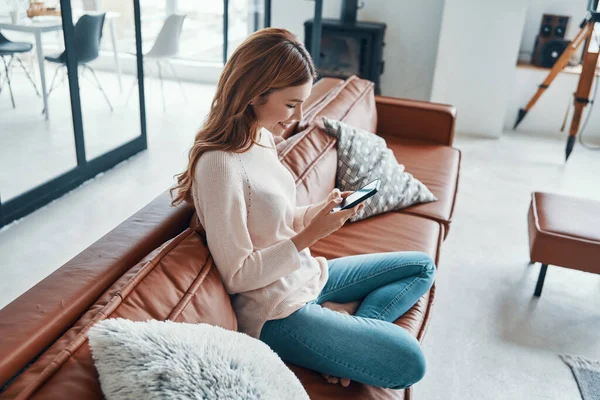 Attractive young woman using smart phone and smiling while spending time at home — Stock Photo, Image