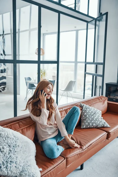 Lächelnde junge Frau am Telefon, während sie zu Hause auf dem Sofa sitzt — Stockfoto