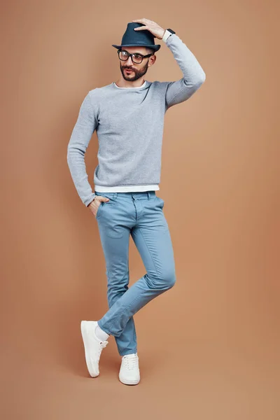 Longitud completa del hombre joven de moda en ropa casual que ajusta el sombrero mientras está parado contra el fondo marrón — Foto de Stock