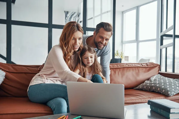 Νεαρή όμορφη οικογένεια με το κοριτσάκι να δένεται και να χαμογελά ενώ χρησιμοποιεί το laptop στο σπίτι — Φωτογραφία Αρχείου
