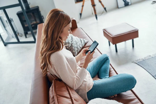 Vacker ung kvinna som använder smart telefon och ler medan du spenderar tid hemma — Stockfoto