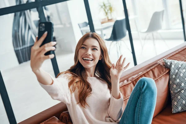 Hermosa mujer joven usando el teléfono inteligente para tomar selfie mientras está sentado en el sofá en casa — Foto de Stock