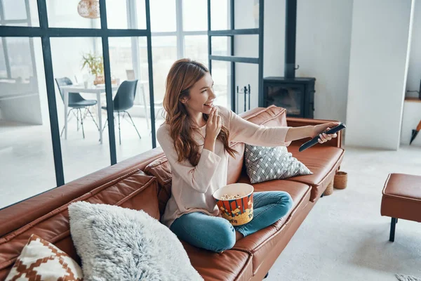 아름다운 젊은 여자가 집의 소파에 앉아 텔레비전을 보고 팝콘을 먹고 있다 — 스톡 사진