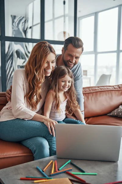 Junge, schöne Familie mit kleinem Mädchen, das zusammenhält und lächelt, während es zu Hause Laptop benutzt — Stockfoto