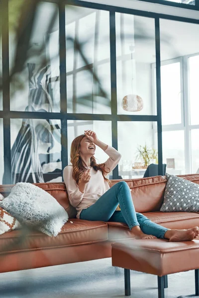 Красивая молодая женщина в повседневной одежде смеется сидя дома на диване — стоковое фото