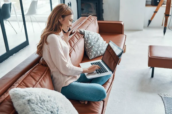 Mujer joven atractiva usando el ordenador portátil y hablando por teléfono mientras pasa tiempo en casa —  Fotos de Stock