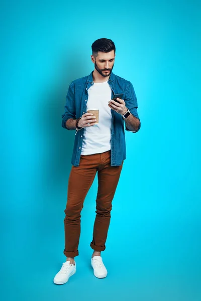 Full length of handsome young man using smart phone while standing against blue background — Stock Photo, Image