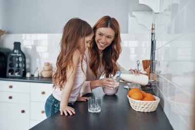 Anne ve kızı mutfakta sütü bardaklara dökerken gülümsüyor.