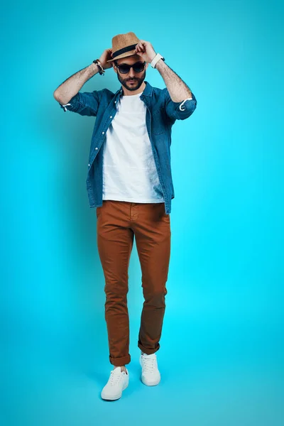 Full length of handsome young man adjusting hat and looking at camera while standing against blue background — Stock Photo, Image