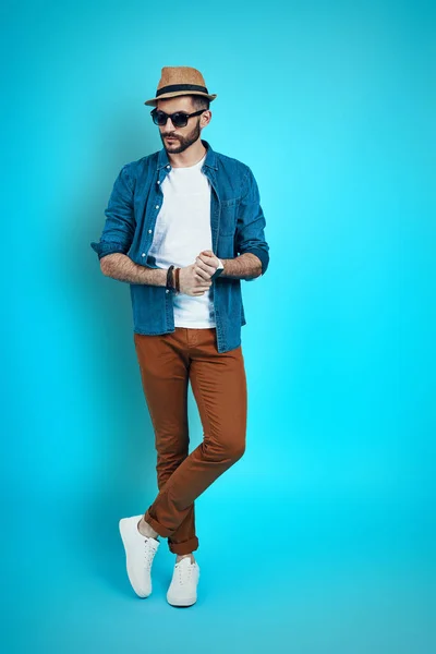 Full length of stylish young man wearing hat and looking away while standing against blue background — Stock Photo, Image