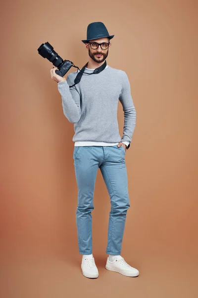 Full längd charmig ung man i casual wear leende och tittar på kameran när du står mot brun bakgrund — Stockfoto