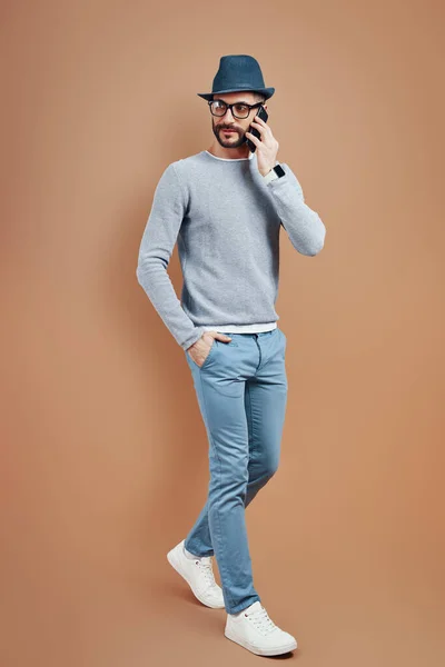Повна довжина молодого чоловіка в повсякденному одязі розмовляє на смартфоні, стоячи на коричневому фоні — стокове фото