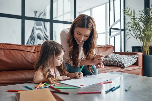 Young beautiful mother and her little daughter drawing in sketchbook while sitting on the sofa ay home — ストック写真