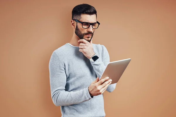 Ragazzo premuroso in abbigliamento casual intelligente tenere la mano sul mento e utilizzando tablet digitale mentre in piedi su sfondo marrone — Foto Stock