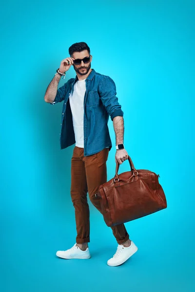 Full length of stylish young man smiling and adjusting eyewear while walking against blue background — Stock Photo, Image