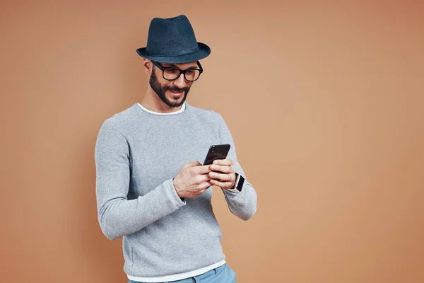 Hombre joven guapo en ropa casual usando el teléfono inteligente mientras está de pie contra el fondo marrón — Foto de Stock