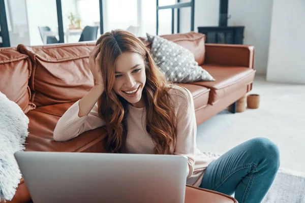 Mujer joven atractiva usando el ordenador portátil y sonriendo mientras pasa tiempo en casa —  Fotos de Stock