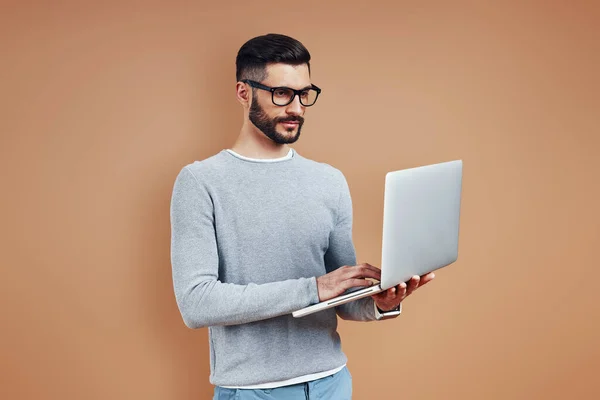 Busy young man in smart casual wear using laptop while standing against brown background — Stock Photo, Image