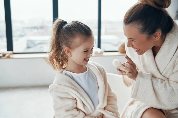 Madre con hija en albornoces sonriendo y usando crema de manos mientras hace la rutina de la mañana —  Fotos de Stock