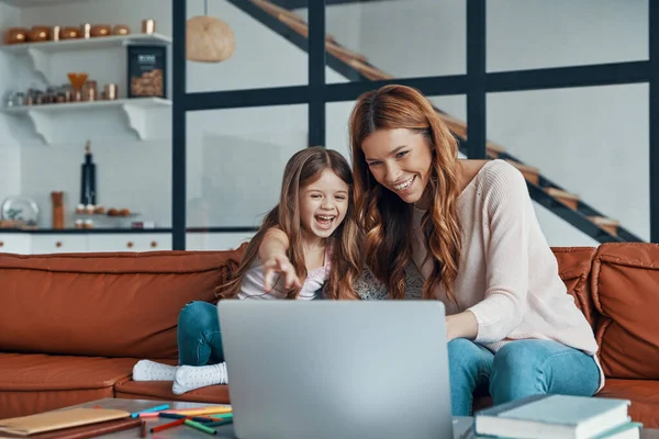 Fiatal gyönyörű anya és kislánya összebarátkoznak és mosolyognak, miközben otthon laptopot használnak. — Stock Fotó
