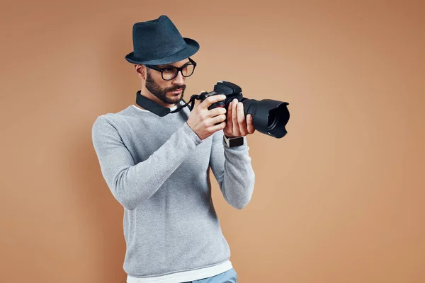 Очаровательный молодой человек в повседневной одежде регулирует фотокамеру, стоя на коричневом фоне — стоковое фото