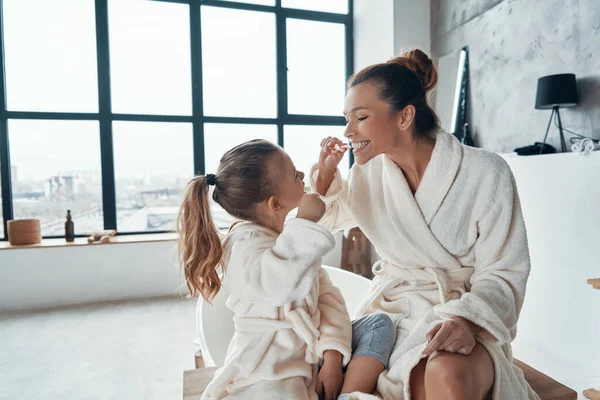 Madre e hija en albornoces sonriendo y limpiando los dientes mientras hacen la rutina de la mañana —  Fotos de Stock