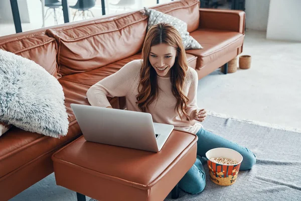 Приваблива молода жінка використовує ноутбук і їсть попкорн, проводячи час вдома — стокове фото
