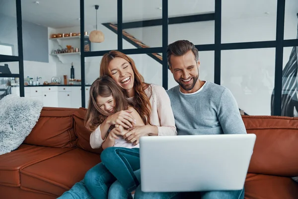 Juguetona hermosa familia con niña que se une y sonríe mientras usa el ordenador portátil en casa —  Fotos de Stock