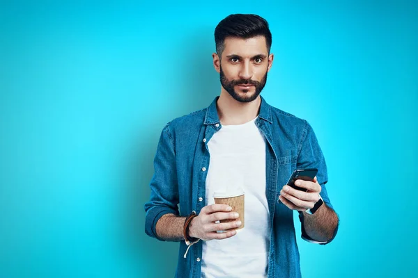 Hombre joven guapo sonriendo y utilizando el teléfono inteligente, mientras que de pie sobre fondo azul — Foto de Stock