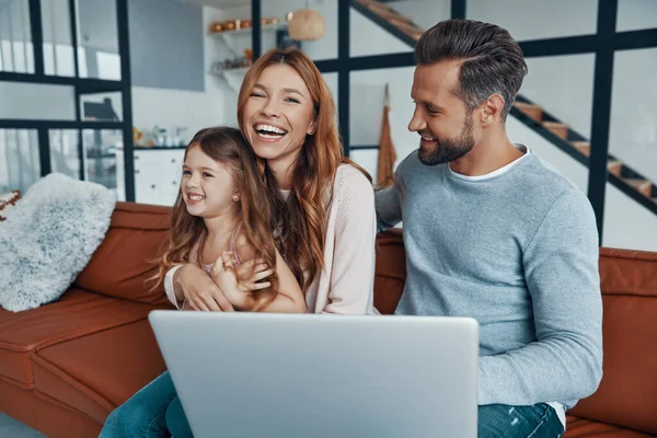 Juguetona hermosa familia con niña que se une y sonríe mientras usa el ordenador portátil en casa —  Fotos de Stock