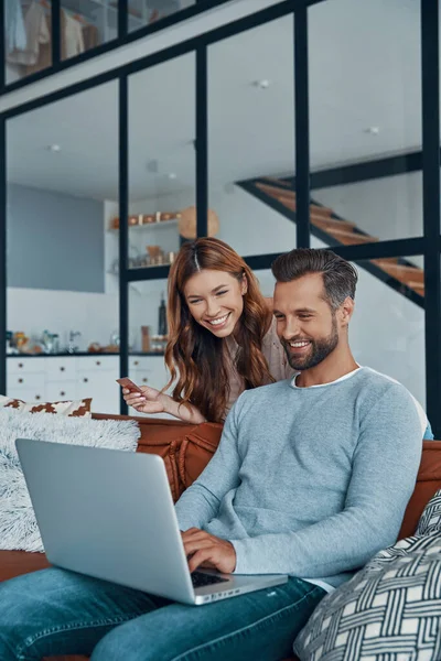 Alegre pareja joven utilizando el ordenador portátil y sonriendo mientras que las compras en línea en casa —  Fotos de Stock