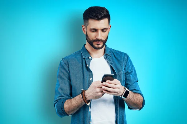 Jovem bonito usando telefone inteligente enquanto está de pé contra o fundo azul — Fotografia de Stock