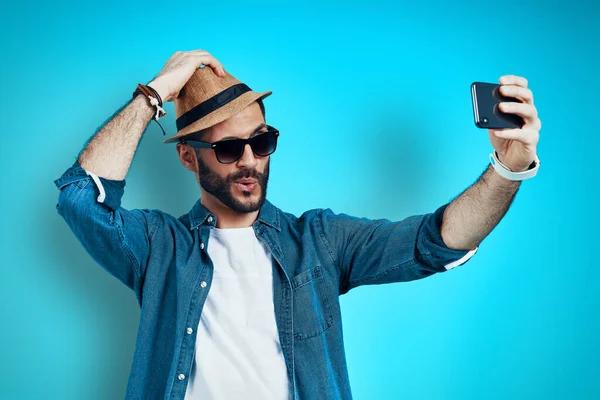 Hombre joven de buen aspecto sonriendo y tomando selfie usando el teléfono mientras está parado contra el fondo azul —  Fotos de Stock