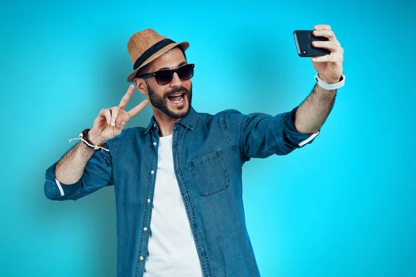 Joven juguetón sonriendo y tomando selfie usando el teléfono mientras está parado contra el fondo azul —  Fotos de Stock