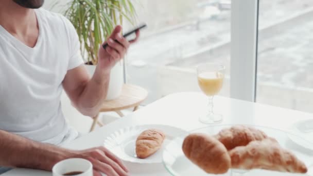 Snygg ung man pratar i telefon medan du äter frukost i köket — Stockvideo