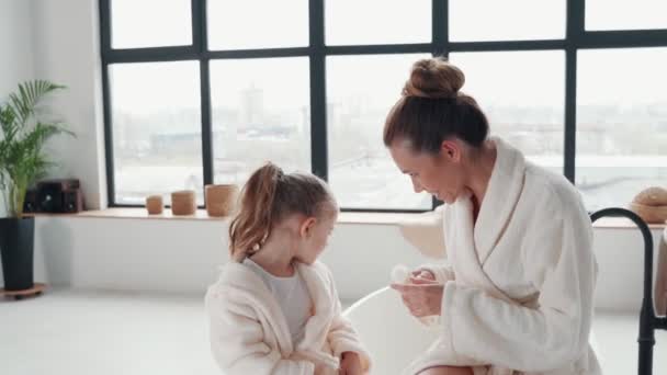 Speelse moeder met dochter in badjassen glimlachen en met behulp van handcrème — Stockvideo