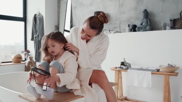 Jovem mãe alegre combinando seu cabelo filha enquanto faz rotina matinal — Vídeo de Stock