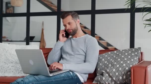 Молодой человек в повседневной одежде, используя ноутбук и разговаривая на смартфоне — стоковое видео