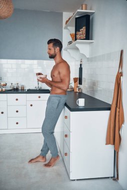 Genç, üstsüz bir adam kahvaltısını yaparken mutfakta dikiliyor..