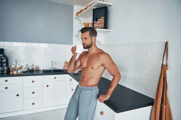 Bello sguardo giovane uomo senza maglietta bere tè caldo mentre in piedi a casa cucina — Foto Stock