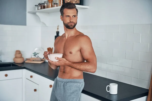 Good looking young shirtless man having breakfast while standing at home kitchen