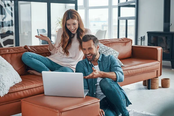 Belo jovem casal usando laptop e sorrindo enquanto passa o tempo em casa — Fotografia de Stock