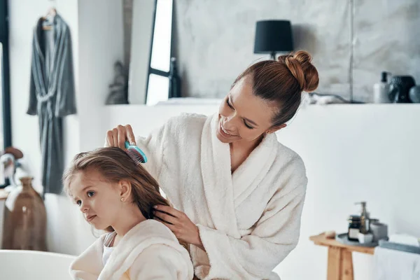 Bela jovem mãe combinando seu cabelo filha ao fazer rotina matinal — Fotografia de Stock