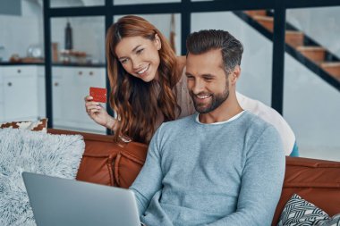 Mutlu genç çift evde online alışveriş yaparken dizüstü bilgisayar kullanıyor ve gülümsüyor.