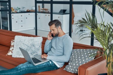 Laptop kullanan ve akıllı telefondan konuşan rahat giyimli yakışıklı bir genç.