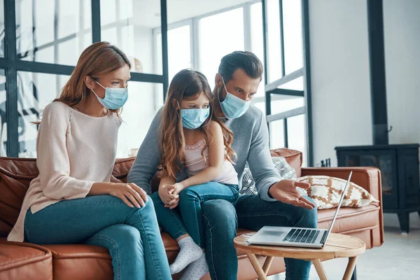 Hermosa familia con mascarillas protectoras y el uso de ordenador portátil en casa —  Fotos de Stock