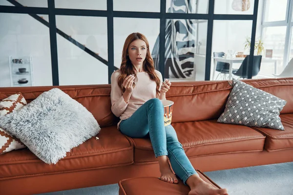 Surprised young woman eating popcorn while sitting on the sofa at home — Stock Photo, Image