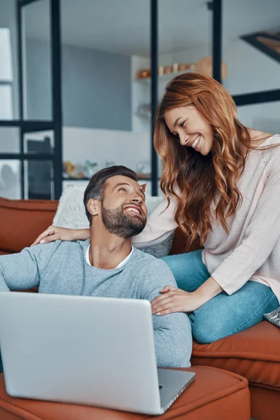 Amar jovem casal usando laptop e sorrindo enquanto passa o tempo em casa — Fotografia de Stock